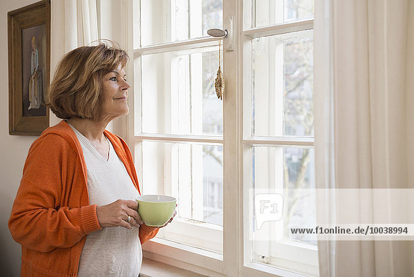 Side profile of a senior woman having cup of tea looking through window at home  Munich  Bavaria  Germany