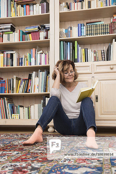 Senior woman sitting on the floor in front of bookshelf and reading  Munich  Bavaria  Germany