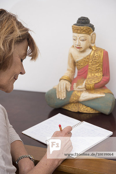 Senior woman writing in notebook  Buddha statue in the background  Munich  Bavaria  Germany