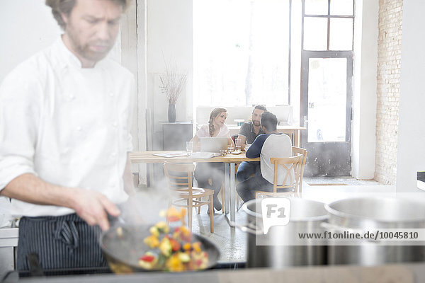 Koch arbeitet in der Küche seines Restaurants  während die Gäste im Hintergrund kommunizieren.