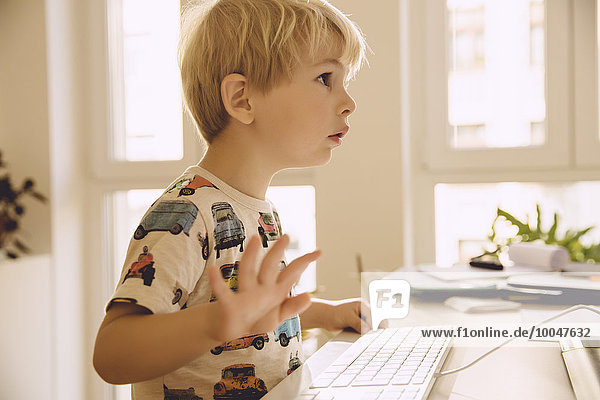 Blonder kleiner Junge  der auf der Tastatur eines Computers tippt und auf den Bildschirm schaut.