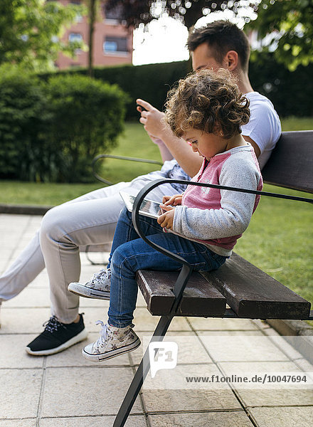 Young man and little girl sitting side by side on a bench looking at smartphone and digital tablet