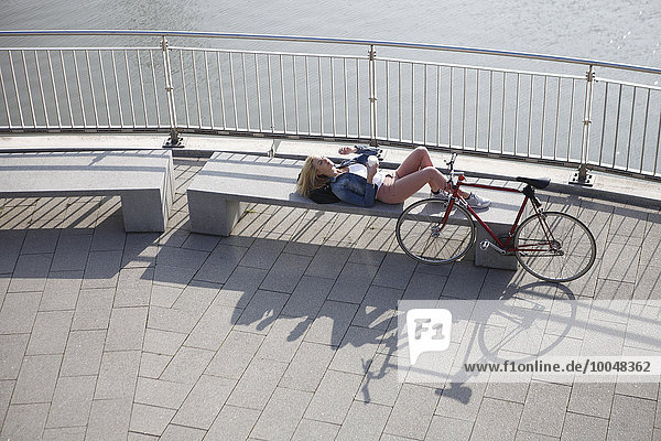 Deutschland  Düsseldorf  blonde Frau liegt auf einer Bank und ruht sich vor dem Rhein aus.