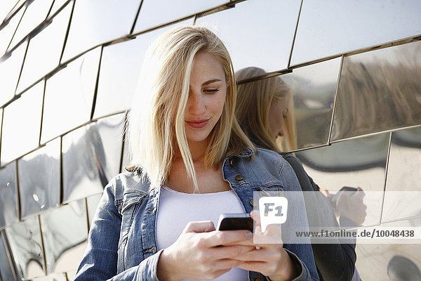 Germany  Duesseldorf  smiling blond woman looking at her smartphone in front of modern facade