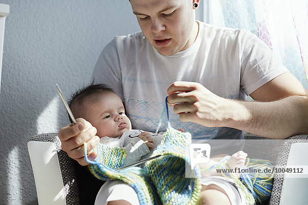 Junger Vater strickt mit Baby auf dem Schoß