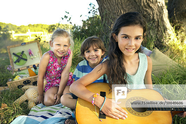Mädchen am Baum hält Gitarre mit Geschwistern im Hintergrund
