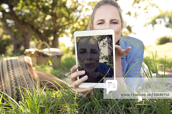Frau mit digitalem Tablett auf der Wiese liegend mit einem Selfie