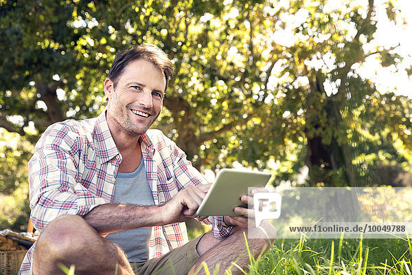 Lächelnder Mann mit digitalem Tablett auf der Wiese sitzend
