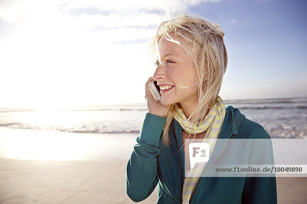 Lächelnde junge Frau am Strand bei Sonnenaufgang per Handy