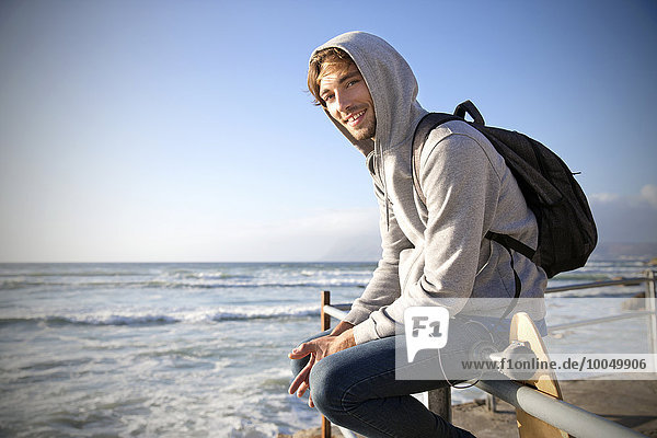 Lächelnder junger Mann sitzt auf einem Geländer an der Küste
