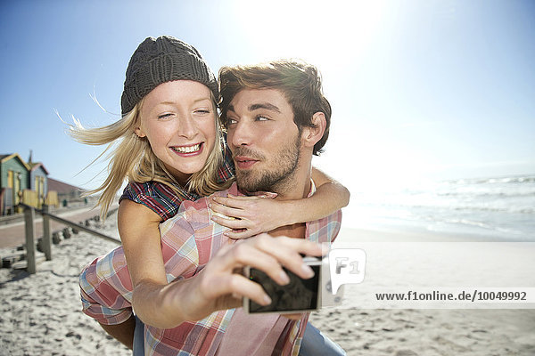 Junge Frau auf dem Rücken ihres Freundes am Strand,  die einen Selfie nimmt.
