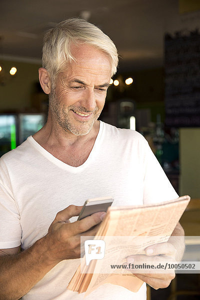 Porträt eines lächelnden Mannes mit Smartphone beim Zeitungslesen