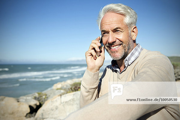 Südafrika  Porträt eines lächelnden Mannes  der auf Felsen sitzt und mit dem Smartphone telefoniert.