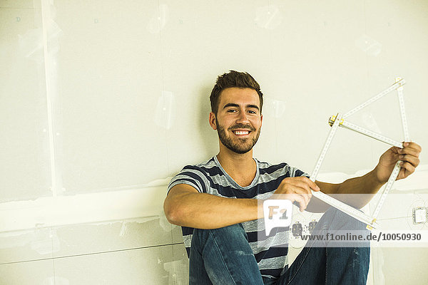 Smiling young man sitting at blank wall holding pocket rule