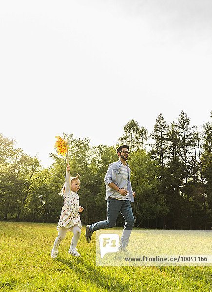 Father and daughter running with paper windmill on meadow