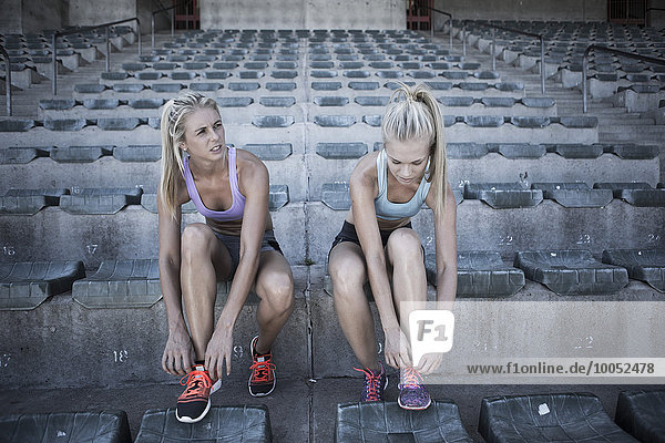 Zwei Sportlerinnen sitzen auf der Tribüne eines Stadions und binden ihre Schuhe.