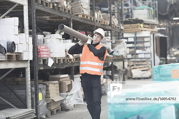 Young male warehouse worker carrying industrial pipe on shoulder