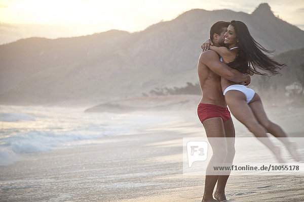 Mittleres erwachsenes Paar am Strand  trägt Badebekleidung  Mann schwingt Frau herum