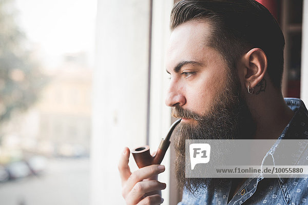 Junger bärtiger Mann raucht Pfeife am Fenster