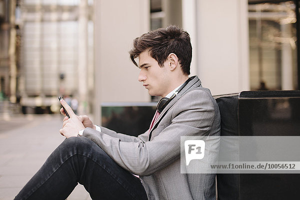 Ein junger Geschäftsmann sitzt auf dem Bürgersteig und liest Smartphone-Nachrichten.