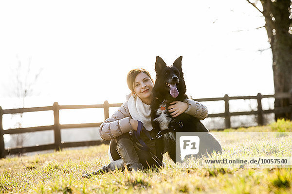 Porträt einer mittleren erwachsenen Frau  die mit ihrem Hund auf dem Feld sitzt.
