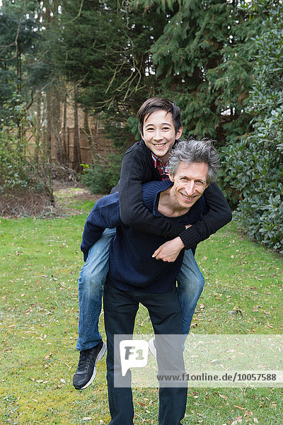 Portrait of mature man giving teenage son a piggy back in garden