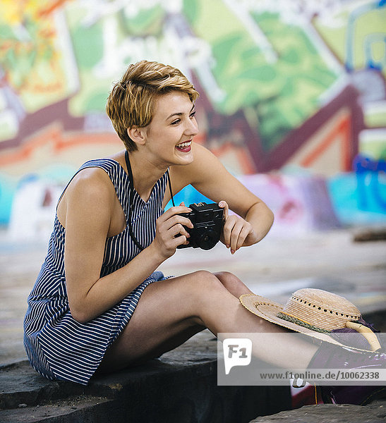 Stilvolles Teenagermädchen auf dem Bürgersteig sitzend mit Kamera vor der Graffiti-Wand