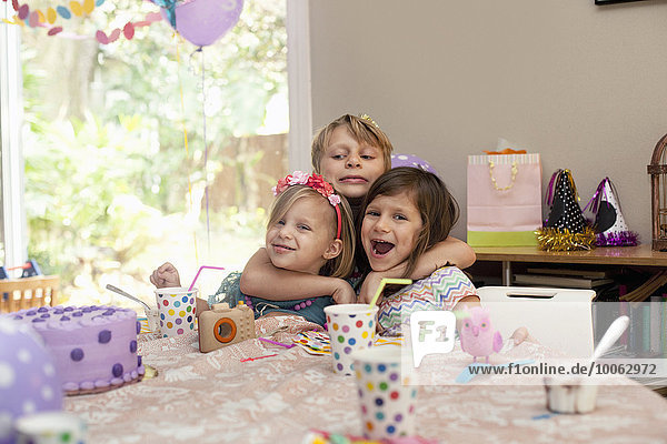 Three children sitting at birthday party table hugging each other