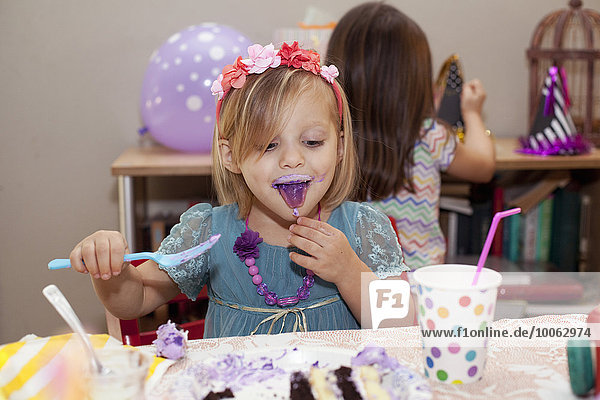 Porträt eines Mädchens  das am Geburtstagstisch sitzt und lila Kuchen mit lila Zunge isst.