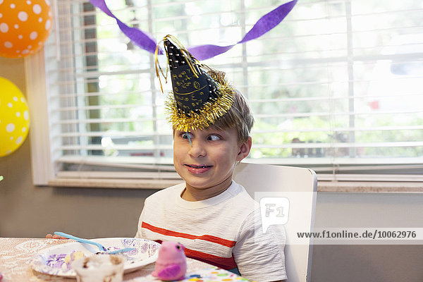 Junge mit Partyhut sitzt am Tisch und isst Geburtstagstorte mit lustigem Gesicht.