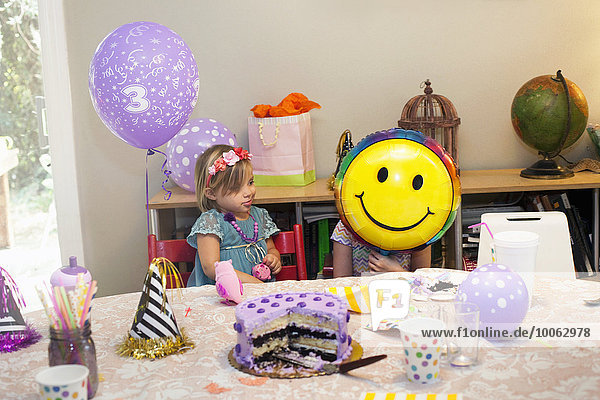 Zwei Mädchen sitzen am Geburtstagstisch und spielen mit einem Smiley-Ballon.