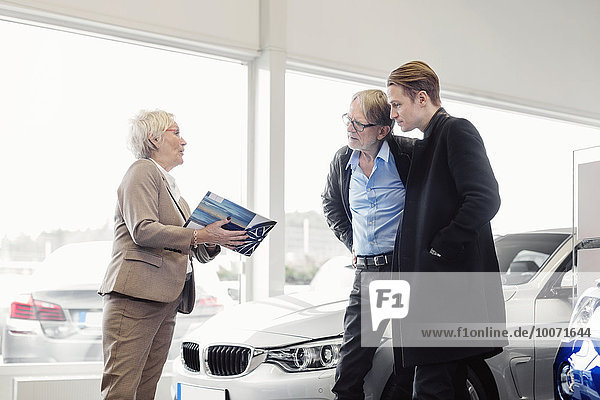 Senior saleswoman explaining brochure to son and father in car dealership shop