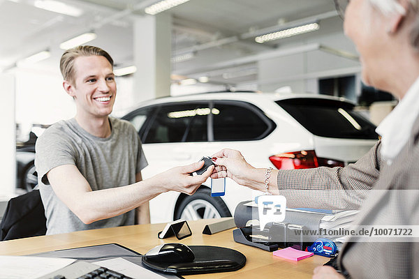Glücklicher Mann erhält Autoschlüssel von Verkäuferin am Schreibtisch im Showroom