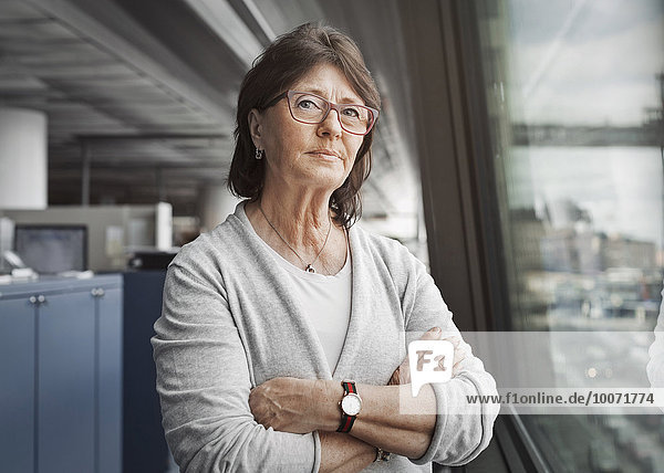 Portrait of confident senior businesswoman standing arms crossed by window in office
