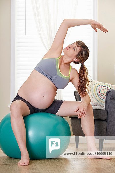 Schwangere Frau beugt sich seitwärts  während sie auf dem Übungsball im Wohnzimmer sitzt.