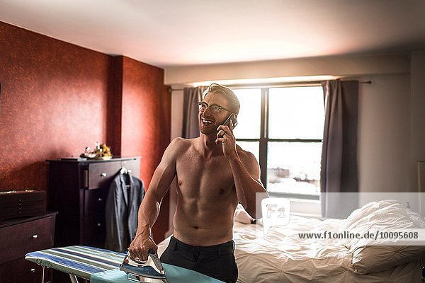 Mid adult man wearing boxer shorts chatting on smartphone whilst ironing