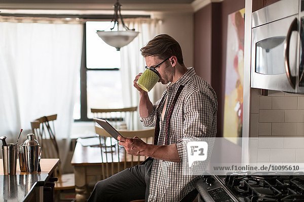 Mid adult man in kitchen drinking coffee whilst reading digital tablet