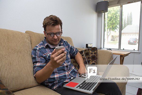 Junger Mann sitzt auf dem Sofa  benutzt den Laptop und schaut auf das Smartphone.