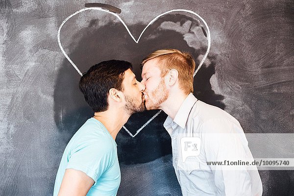 Männliches Paar  das sich vor einer Tafel mit Kreideherz küsst.