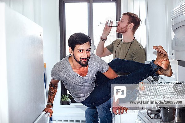 Male couple at home  mid adult man drinking whilst his partner balances on kitchen appliances