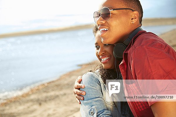 Junges Paar steht zusammen am Strand und lächelt  umarmt und schaut weg.