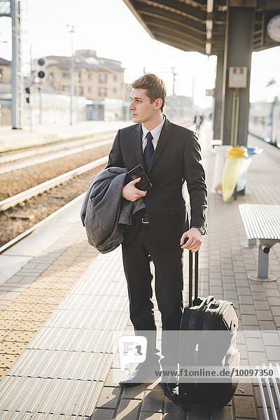 Young businessman commuter standing on railway platform with suitcase.