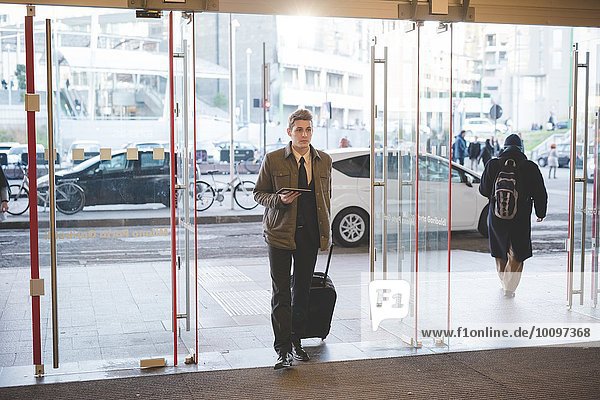Young businessman commuter walking through glass doors with suitcase.