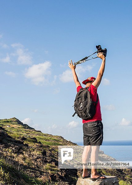 Junger männlicher Tourist  der seine digitale Spiegelreflexkamera auf dem Küstenweg von Makapuuu  Oahu  Hawaii  USA  hochhält.