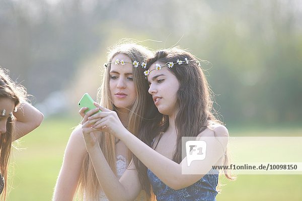 Zwei Teenager-Mädchen in Gänseblümchen-Kopfbedeckungen beim Lesen von Smartphone-Texten im Park