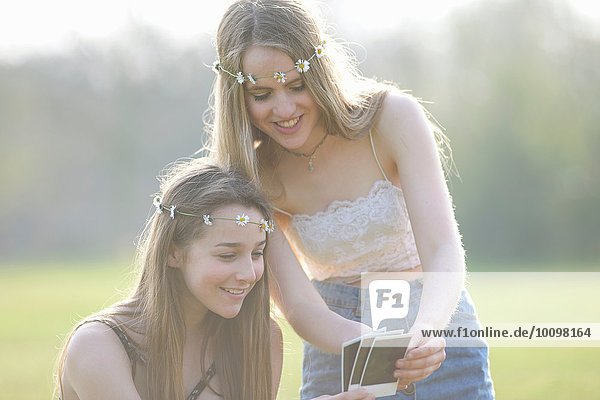 Zwei Teenager-Mädchen in Gänseblümchen-Kopfbedeckungen  die sich Sofortbilder im Park ansehen.