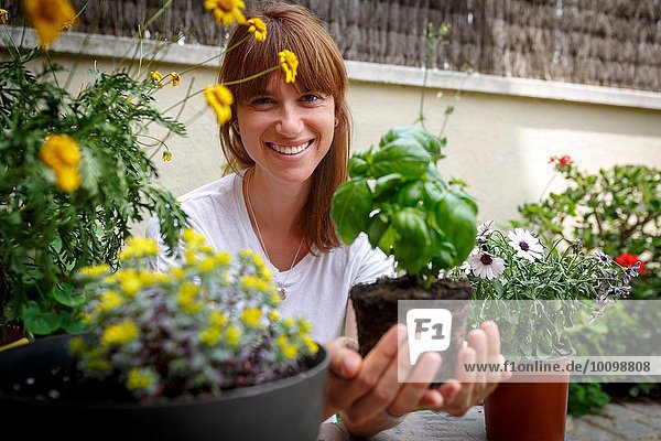 Mittlere erwachsene Frau hält Basilikumpflanze in hohlen Händen  lächelnd vor der Kamera