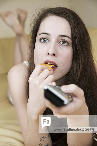 Junge Frau sieht Fernsehen auf dem Sofa mit Chips und der Fernbedienung in der Hand