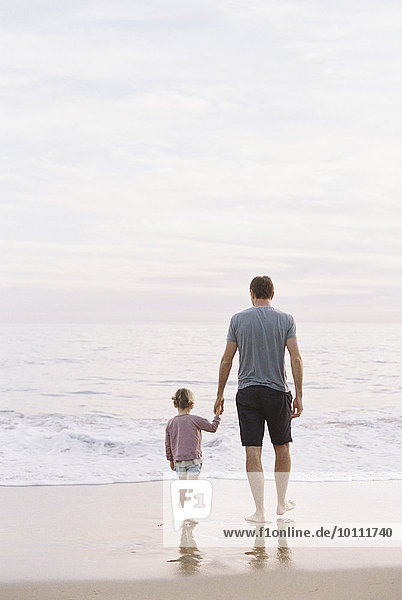 Man standing on a sandy beach by the ocean  holding his young daughter's hand.