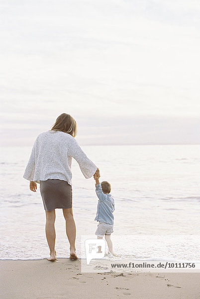 Woman standing on a sandy beach by the ocean  holding her young son's hand.